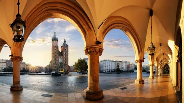 Krakau Geheimtipps: Blick durch Arkaden auf den Marktplatz mit Marienkirche im Sonnenaufgang. Ein malerischer Ort für Entdeckungen.
